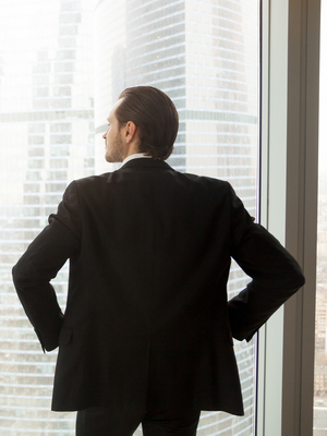 Back view of man in suit standing with hands on hips looking on cityscape outside the window. Successful businessman thinking about future plans, guy dreams about great career. Ambitions in business